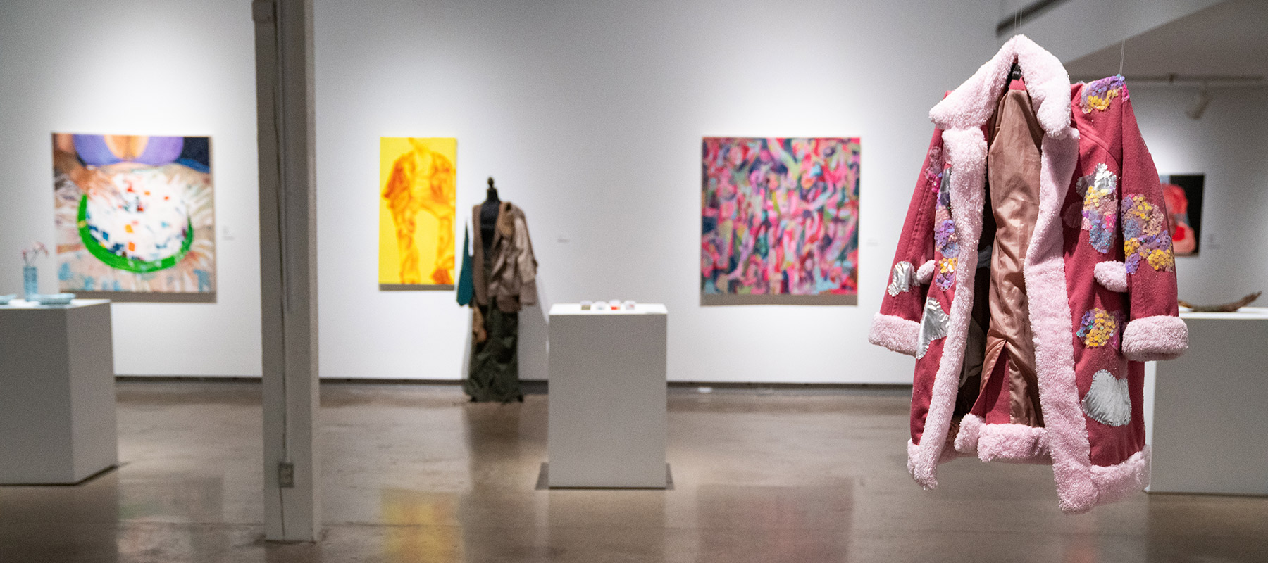a pink fluffy overcoat with patches of sequins sewn onto it, suspended in front of several paintings on the wall, three pedestals holding artwork, and a headless mannequin wearing a patchwork suit