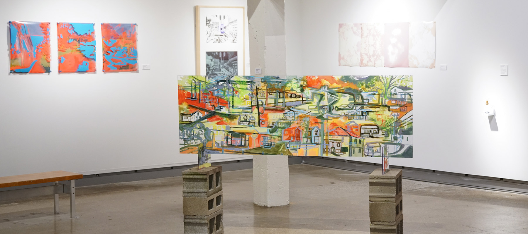 an abstract painting of a town full of houses, roads, and trees on plexiglass resting on stacks of cinderblocks; several artworks mounted on the wall behind it