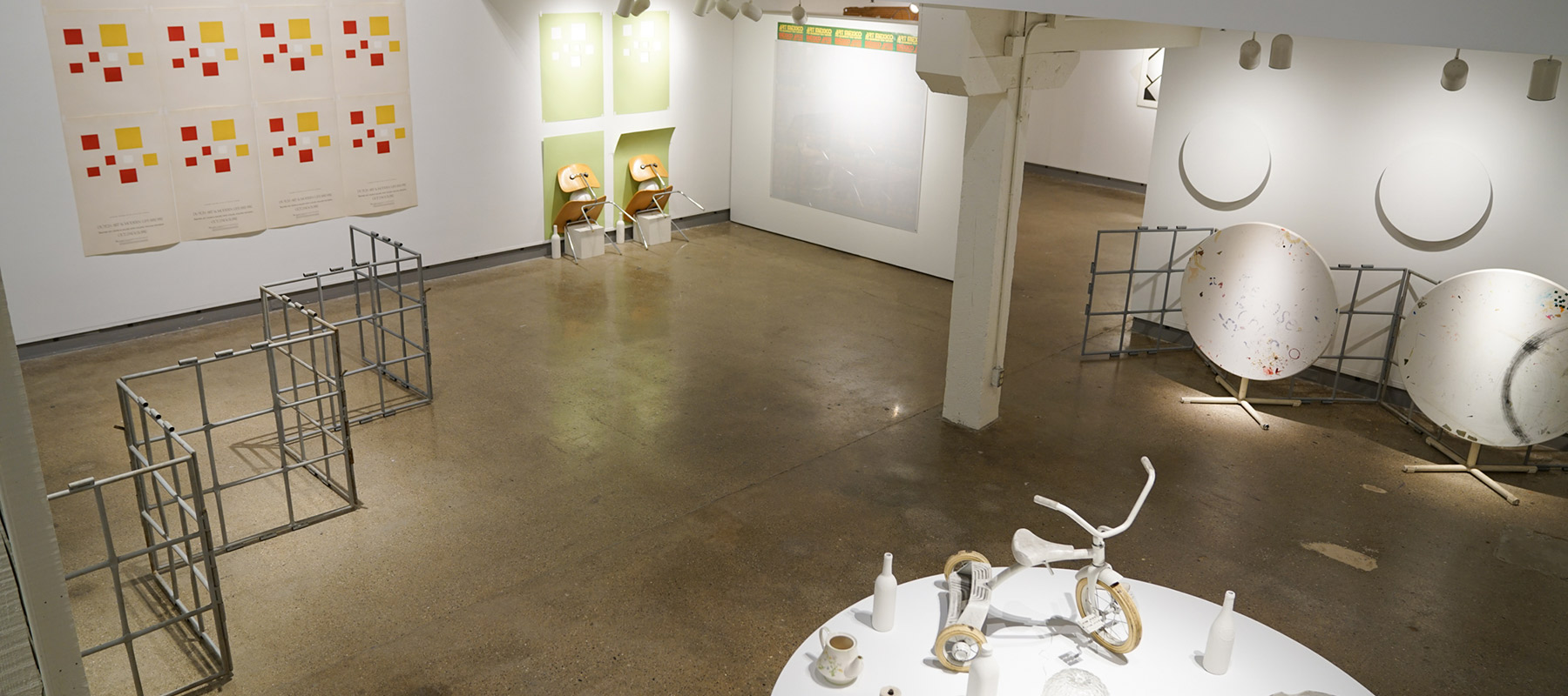 gallery space with posters of red and yellow squares on the wall; metal scaffolding zig-zagged on the floor; and a low podium holding various white objects, including a tricycle and several dishes and cups