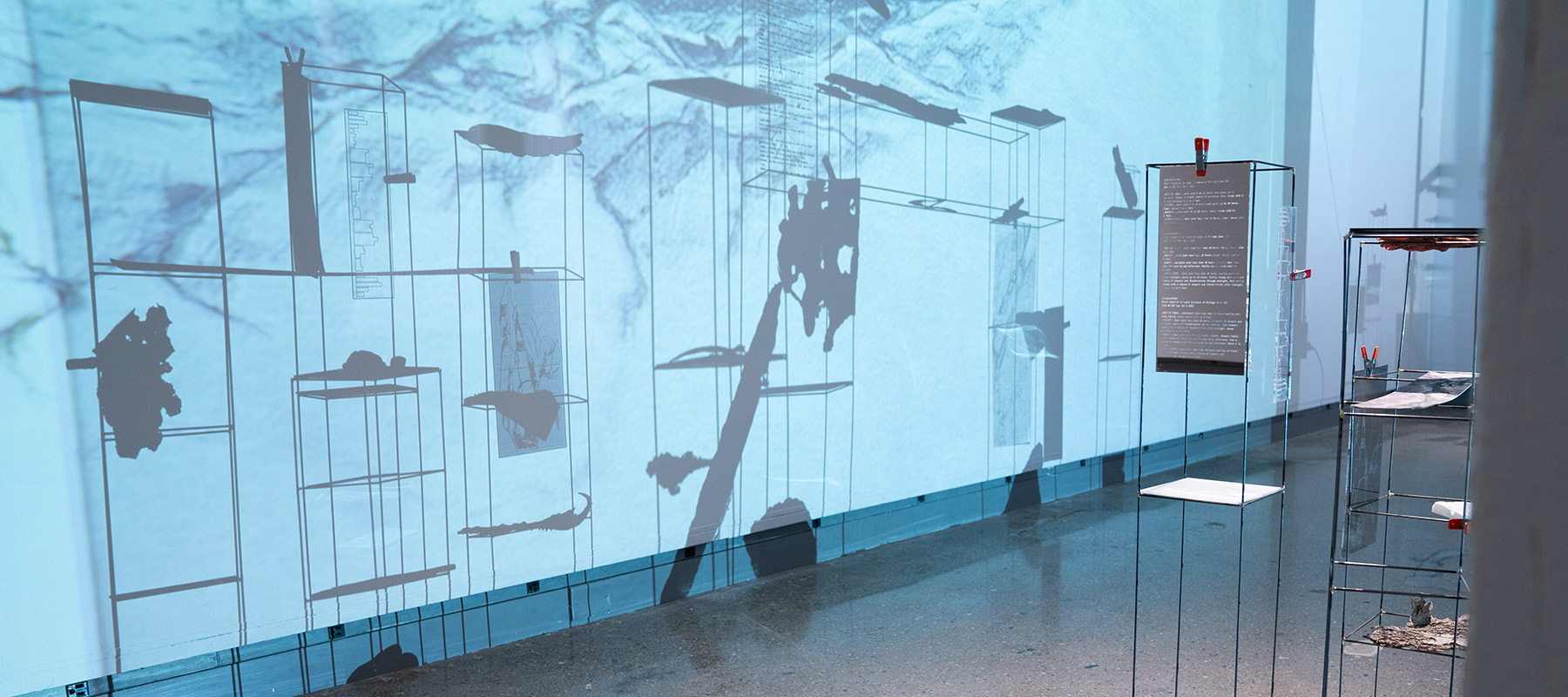 close up image of a rock projected over rectangular wireframe structures with rocks and plexiglass plates clamped to them, casting a shadow of the structures on the wall within the projected image