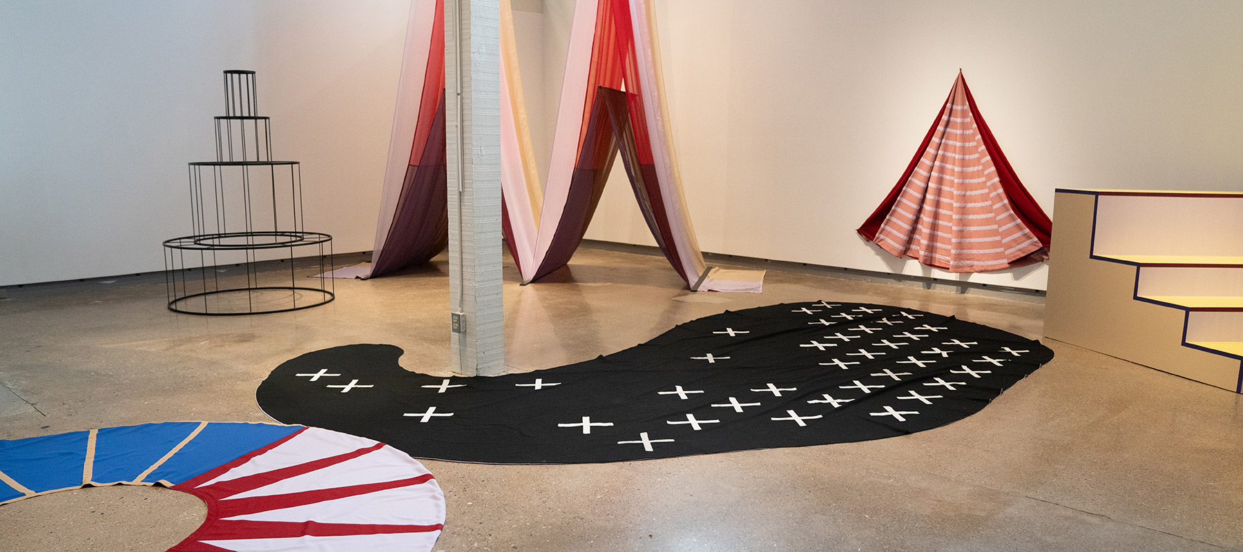 gallery room filled with fabric-based installation artwork, including a large black oblong sheet with white plus signs on the floor; a red and blue ring-shaped sheet on the floor; a wire frame structure that looks like a large round tiered cake; red fabric arranged in a triangle on the wall; layered red and pink sheer fabric that is draped from the ceiling to look like a capital M; and two sets of low wooden stairs painted light yellow