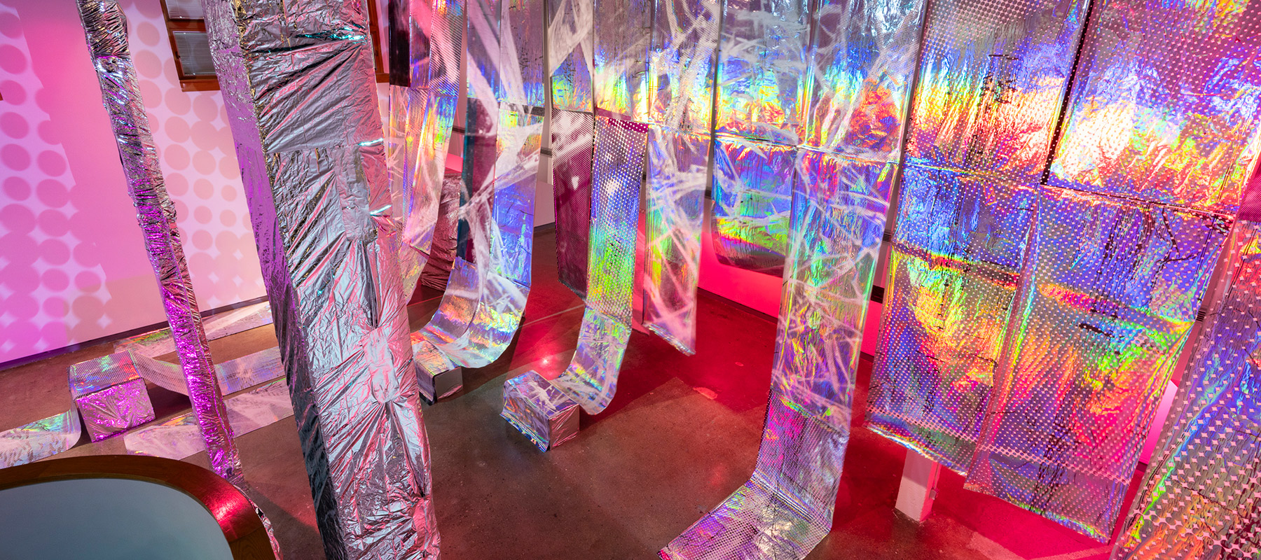 pink light and a halftone dot pattern projected over several long swathes of holographic material draped down from the ceiling